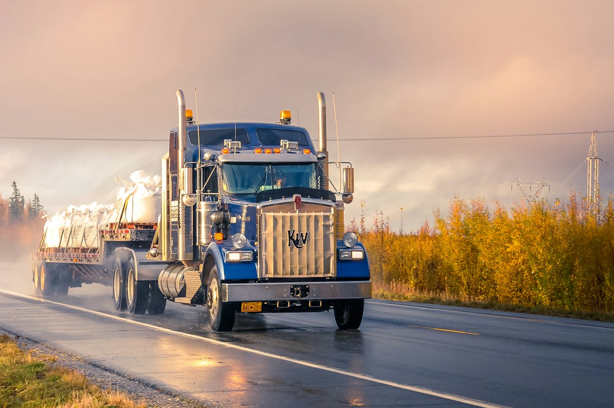 Long-nose semi truck hauling a flatbed trailer on a wet road at sunset